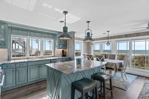Coastal kitchen with blue cabinets, granite island, bar stools, pendant lights, dining table, wide windows, ocean views, and direct beachfront scenery.