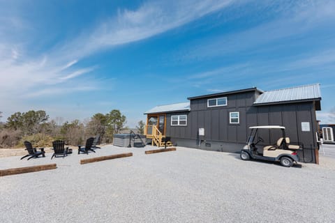 Gravel patio area with black Adirondack chairs, hot tub, and beige golf cart beside dark gray cottage.
