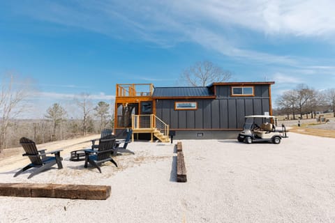 Black Adirondack chairs surround a metal fire pit on gravel beside the charcoal gray cottage with wood trim and rooftop deck.
