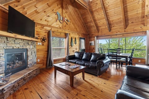 Cozy cabin living room with a stone fireplace, vaulted wood ceilings, and big windows opening to peaceful forest views.