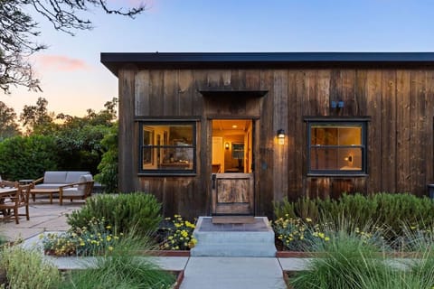 The 1905 farmhouse at dusk — reclaimed barnwood, a Dutch door, and the kitchen glowing from inside.