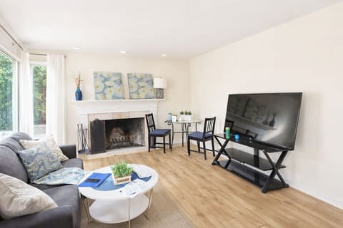 This inviting living area features a charming fireplace and a large flat-screen TV, creating a cozy and functional space for relaxation or movie nights. Modern wood flooring and neutral tones are complemented by stylish blue accents and a secondary bistro