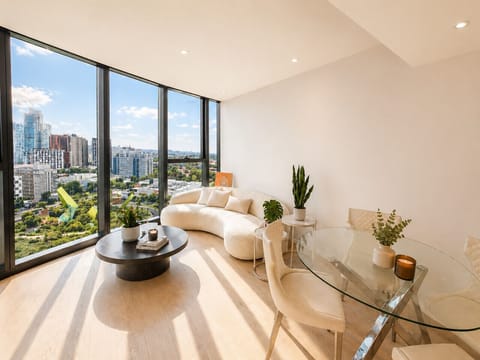 Light-filled living and dining area with floor-to-ceiling windows framing sweeping city and parkland views, featuring a curved bouclé sofa, glass dining table, and warm timber flooring throughout.