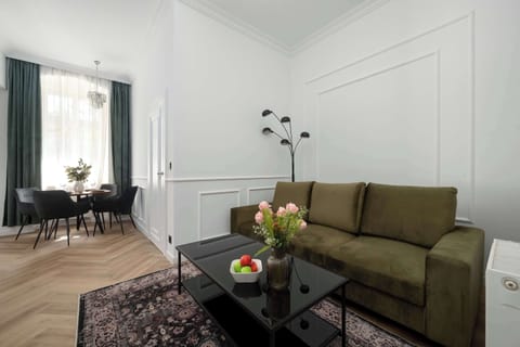 A bright living room with a green sofa, a coffee table, and a TV area, connected to a dining space with a table and chairs. Large windows let in natural light, making the interior feel open and inviting.