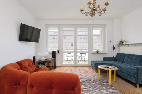 A cozy lounge with an orange sofa facing a TV and a small coffee table. A blue sofa is visible in the background.