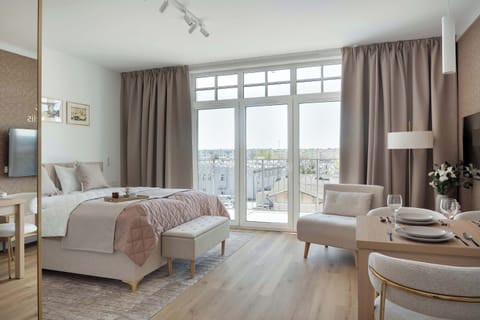 A bright, modern studio apartment features a double bed with neutral linens and a separate dining area. Large glass balcony doors fill the space with light and offer an expansive view.