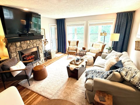 Bright, cozy living room with stone fireplace, big TV, and sunny seating area