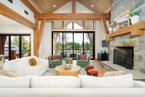 Bright and airy great room with vaulted ceilings, cozy seating, a stone fireplace, and panoramic lake and forest views through glass doors.