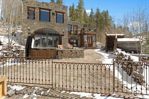 A unique stone lodge nestled among towering pines, featuring a private hot tub on the patio, rustic charm, and ample outdoor space perfect for a mountain escape.