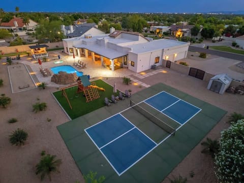 Aerial view of an expansive vacation property featuring a swimming pool, tennis court, playground, and covered patio area in a spacious residential neighborhood at twilight.