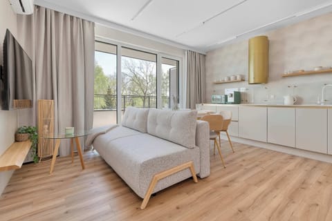 A bright living area featuring a light gray sofa with wooden legs, positioned on warm wood-plank flooring. The space opens up to a large window with floor-to-ceiling curtains, letting in plenty of natural light.