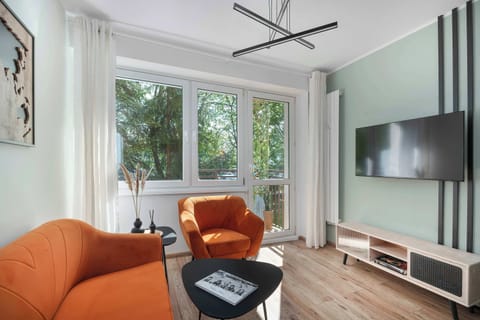 A modern living room features a bright orange sofa and armchair set against a minimalist white wall with a large TV.