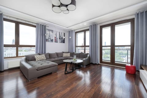 A spacious living room features dark wood-plank flooring and a large, gray sofa. High windows with blue curtains offer a wide view of the city, filling the room with natural light.