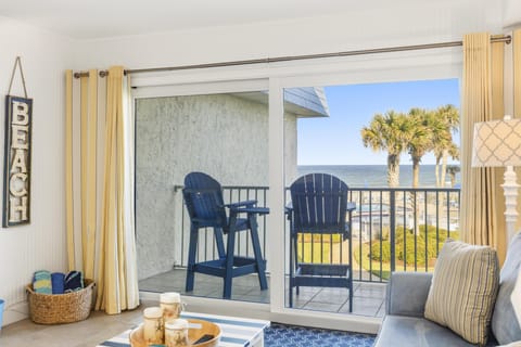 Sliding glass doors open to a balcony with navy Adirondack chairs overlooking palm trees and ocean views.