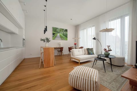 A bright, open-plan living area combining a comfortable sofa, dining space, and wooden flooring with a modern, airy feel. Large windows bring in plenty of natural light and highlight the clean, Scandinavian-style interior.