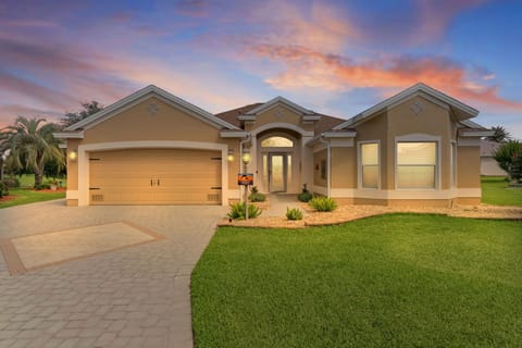 This elegant single-story home features a spacious layout with an arched entrance, welcoming palm trees, and beautifully landscaped grounds under a stunning pastel sunset sky.