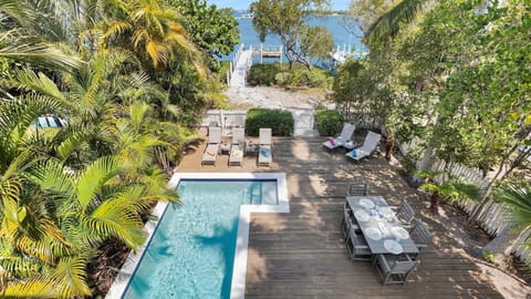 Aerial view reveals a tropical paradise featuring a private pool, waterfront dock access, and expansive deck dining nestled among lush palms and coastal greenery.