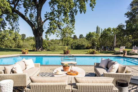 The property features a sparkling outdoor pool surrounded by lush gardens and mature trees, with multiple seating areas for relaxation and socializing in a serene countryside setting.