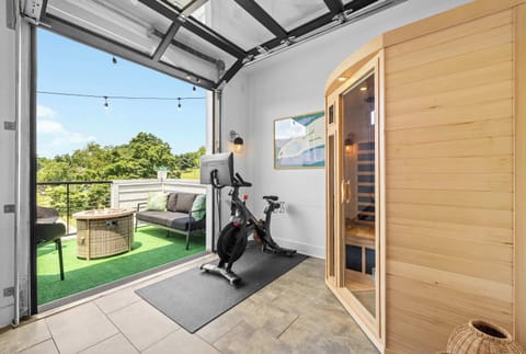 The Wellness Studio features a Peloton bike, infrared sauna, yoga station with mat and blocks, and a wall-mounted TV — all inside a bright, airy space with a full garage door that opens to the outdoors.