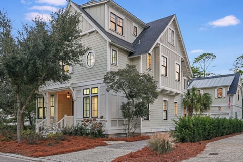 A spacious coastal-style home with classic architectural details, mature landscaping, and warm lighting that welcomes guests at twilight.
