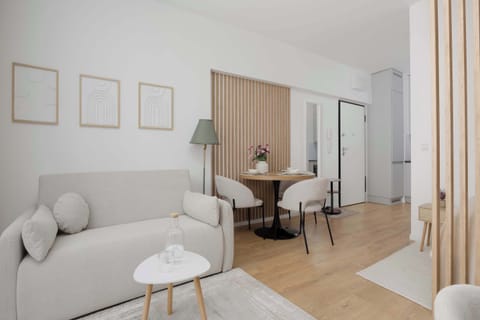 A bright studio apartment with a beige sofa, small dining table, and wooden accent wall beside the bed area. Soft neutral colors and large curtains create a cozy atmosphere.