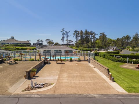 A spacious property featuring a pool, manicured lawns, picnic areas, and mature pine trees, set beneath clear blue skies in a tranquil coastal setting.