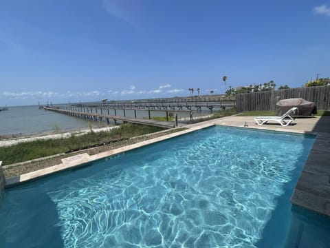 A waterfront pool area overlooking Copano Bay. A long wooden pier extends across the water in the background, with lounge chairs, a privacy fence, and bright blue sky creating a relaxed vacation setting.
