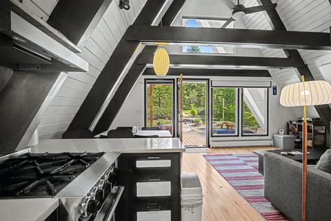 Sunlight streams through skylights onto your open A-frame living space, where modern comforts meet rustic charm. Cook on the sleek gas range while your guests relax on the sofa, all framed by dramatic beams and views of the forest deck beyond.