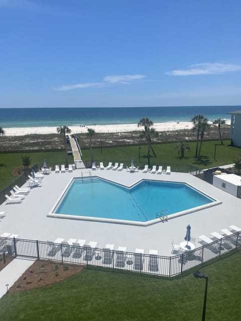 Oceanfront pool area with white lounge chairs, paved deck, and a wooden walkway to the beach under clear blue skies.