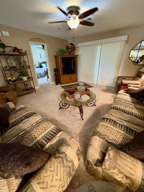 Living room with patterned fabric sofas, glass coffee table on beige carpet, and sliding vertical blinds.
