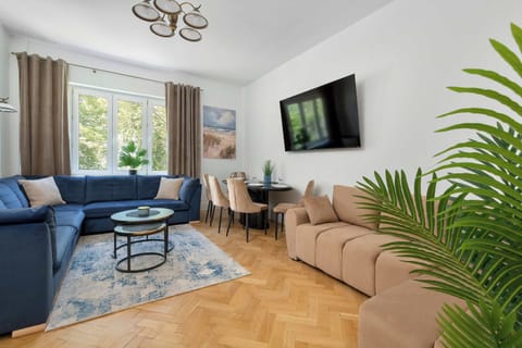 A bright living room features a deep blue sofa, a wall-mounted television, and large windows allowing natural light to fill the space. A decorative rug and indoor plant add warmth to the interior.
