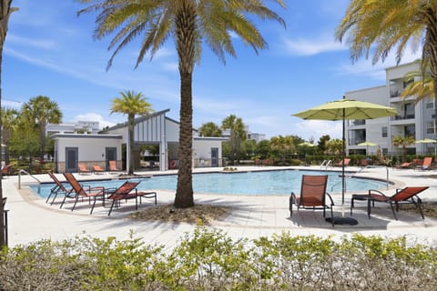 Palm trees surround a kidney-shaped pool with orange mesh loungers and chartreuse umbrellas on white tile decking.