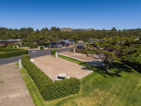 The property features landscaped grounds with manicured lawns, mature evergreen trees, and convenient parking, all surrounded by coastal forestland under clear blue skies.