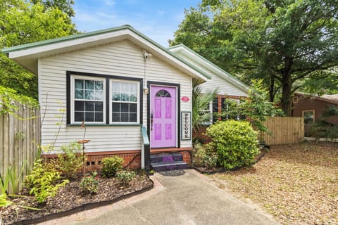 A charming white cottage with a green roof and a bold pink front door, nestled among lush mature trees under a clear blue sky.