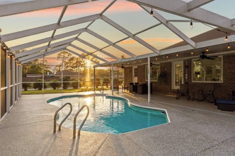 Stunning screened pool enclosure at golden hour, with warm sunset light flooding through the white aluminum frame. A freeform pool with chrome handrails, aggregate deck, and covered patio beyond create a breathtaking private retreat.