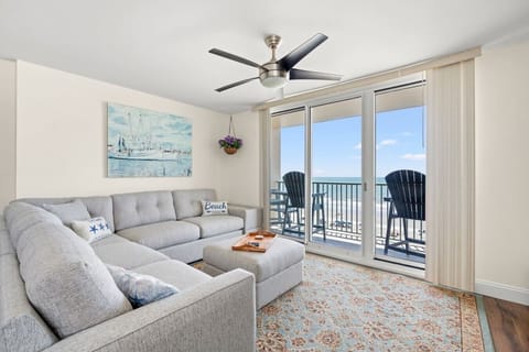 Oceanfront living room at The Pinnacle North Myrtle Beach Unit 506 features a wraparound couch, recliner, large Smart TV, floor-to-ceiling windows, and direct balcony access with stunning Atlantic Ocean views.