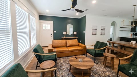 Photo 2 – Living Room  `A stylish living room with a cognac leather sofa, forest green accent chairs, and floor-to-ceiling windows. Bright, welcoming, and designed to feel like home.`