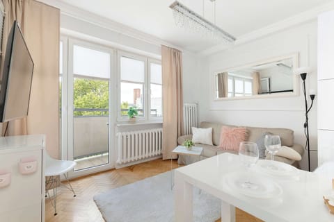 A bright living room with a sofa, coffee table, and large windows overlooking greenery. Soft pink accents and white furnishings create a cozy, inviting atmosphere.