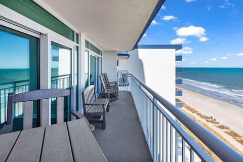Metal railing frames the balcony with gray Adirondack chairs facing the ocean and sandy beach below.