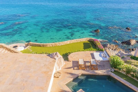 An aerial view showcasing the property's coastal setting with a private pool overlooking pristine turquoise waters and rocky shoreline, surrounded by lush landscaped grounds.
