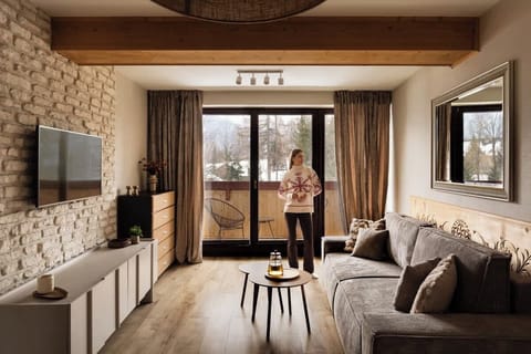 Przytulny salon w górskim stylu z drewnianymi akcentami, kamienną ścianą i wygodną sofą. Duże przeszklenia oraz wyjście na balkon zapewniają piękne widoki i naturalne światło, tworząc idealną przestrzeń do relaksu.