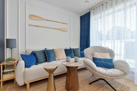 A bright living room features a beige sofa adorned with blue and gold pillows. Wooden accents, a stylish armchair, and decorative oars on the wall create a cohesive coastal ambiance.