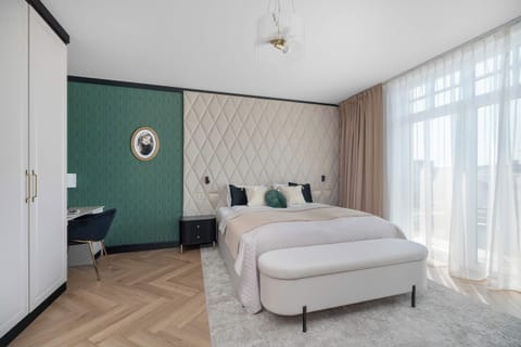 An elegant studio features a bed with a tufted headboard against green wallpaper. An upholstered bench sits on a plush rug by large windows.