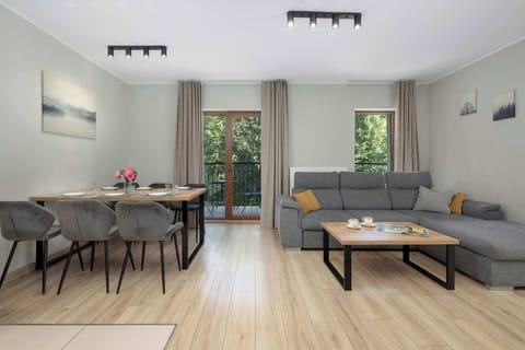 A bright living room with a gray corner sofa, a coffee table, and a dining area. Large windows fill the space with natural light.