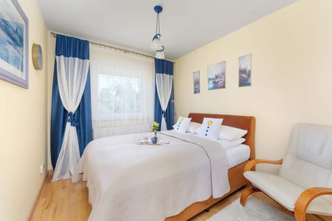 A bright bedroom with a double bed, blue curtains, and light wooden furniture. Large windows bring natural light into the cozy space.
