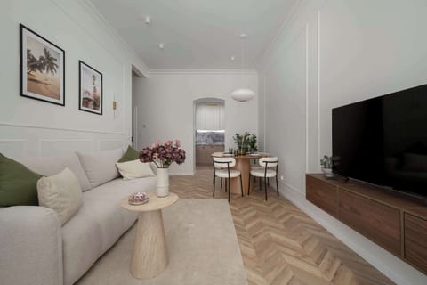 A long view of a modern living room features a light beige sofa on the left and a wall-mounted TV console on the right. Beautiful herringbone wooden floors run throughout the space, leading toward a small dining area in the background.