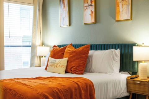 Bright bedroom with a plush bed, crisp white linens, warm orange accents, bedside lighting, and tall windows that bring in natural light for a comfortable and inviting stay.