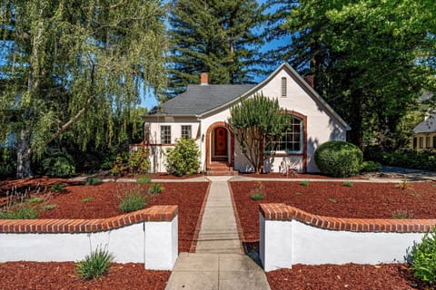 This charming property features a white stucco cottage with red brick accents, nestled among towering evergreens and lush landscaping. The welcoming entrance pathway leads through meticulously maintained gardens with red mulch and manicured shrubs.
