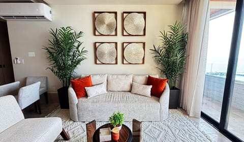 Light beige sofa with red and patterned pillows sits between tall potted plants and textured wall art, near glass doors.