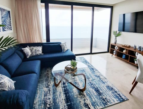 Living room with navy sectional, glass-top coffee table, patterned area rug, and sea view through sliding doors.